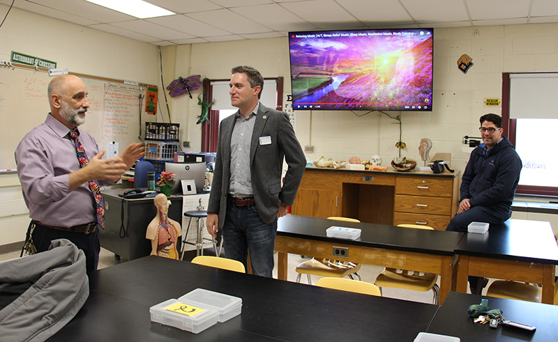 A man on the left talks to another man in the center. Another man sits off to the right.