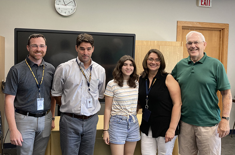 In the center of five people is a high school girl in shorts and shoulder-length brown hair. Next to her are three men and a woman. All are smiling.