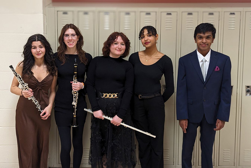 A group of five high school students stand holding various instruments. All are dressed in black dresses and suits.