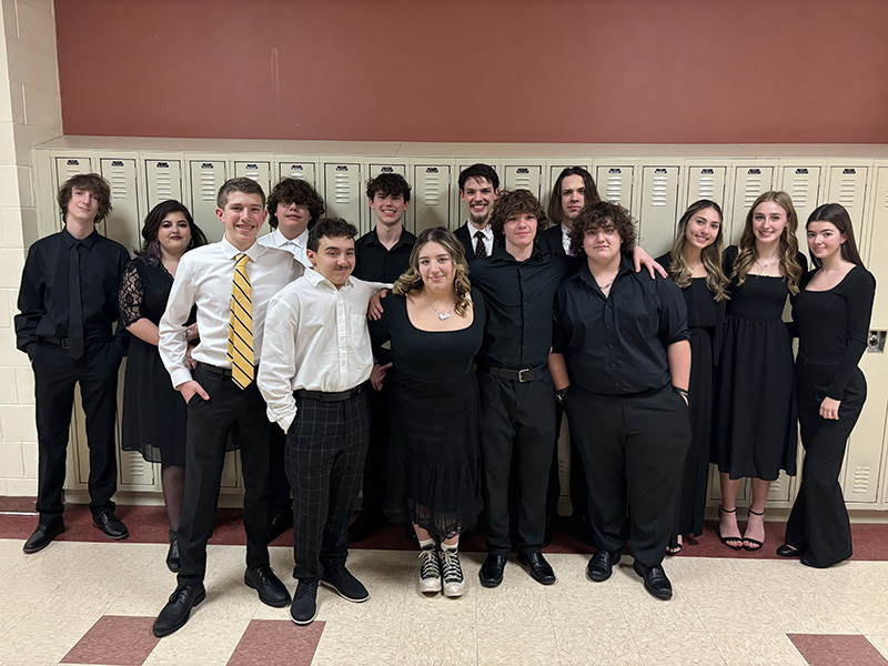 A group of 14 high school students stand together. They are dressed in black dresses and suits, som ewith white shirts and ties.