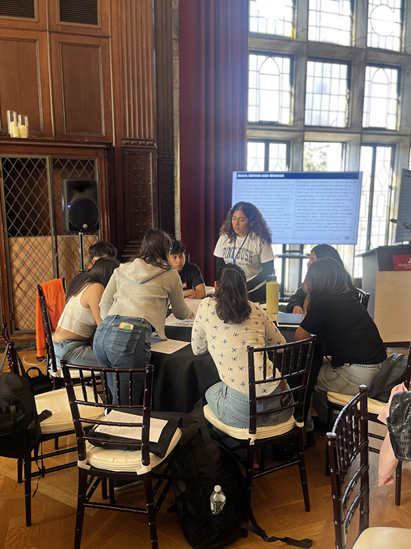 A group of six high school students sit around a table and one stands speaking to them.