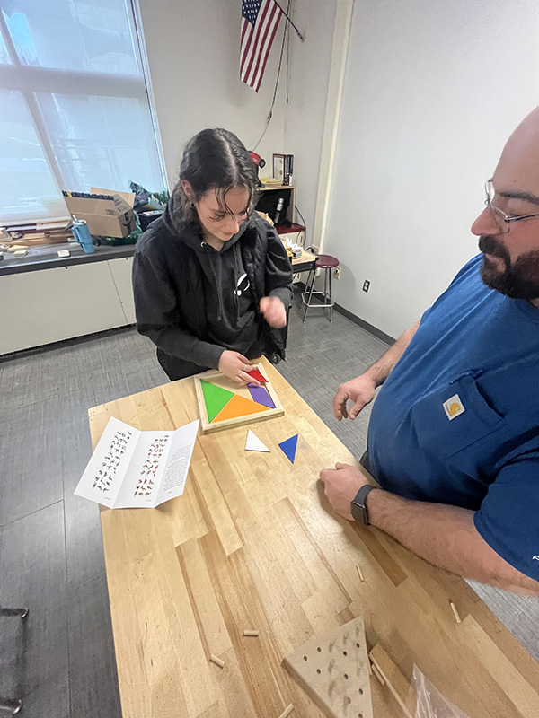 A high school girl works on a colorful wooden puzzle he is making. A man on the right stands and watches.