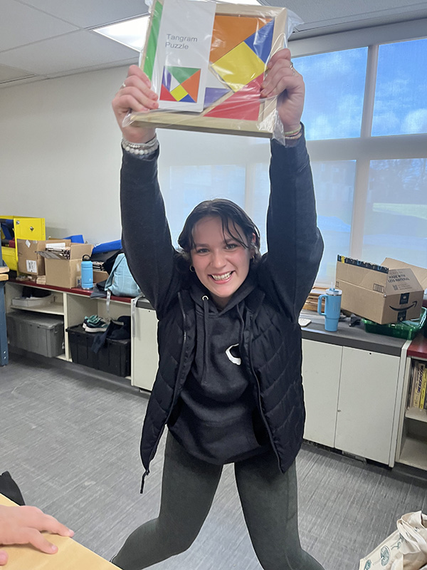 A high school girl with dark hair pulled back holds up over her head a wooden puzzle.