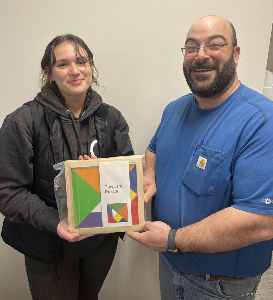 A high school girl on the left with dark hair pulled back holds a wooden puzzle with a man on the right with a beard, wearing a blue shirt.