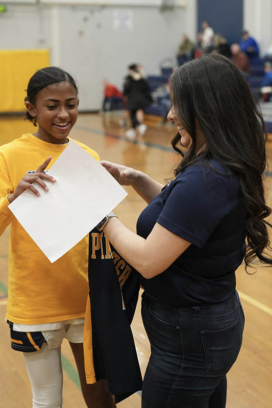 A young basketball player stands on the left handing her basketball jersey to a woman on the right. The woman has long dark hair.