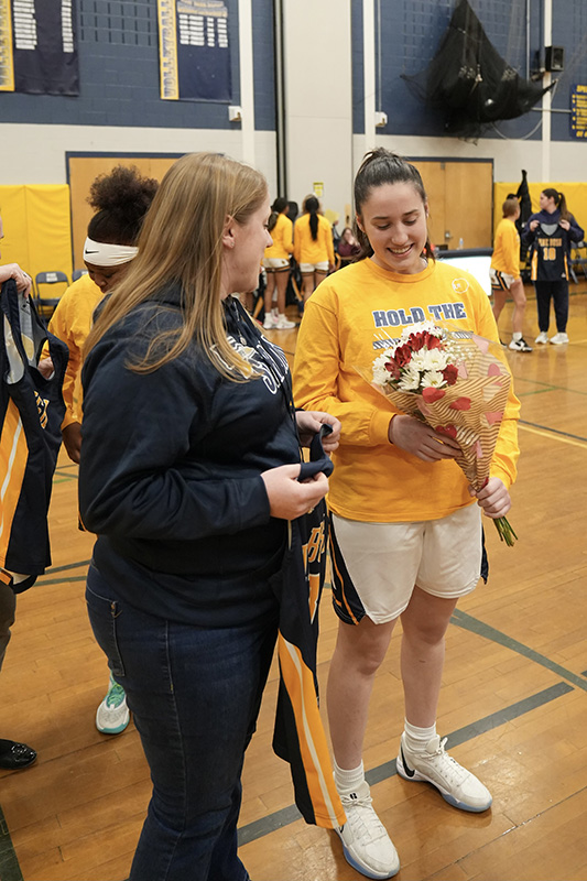 A woman with long blonde hair stands on the left looking at a high school girl on the right who is holding a small bouquet of flowers. The woman is holding a navy blue basketball jersey.