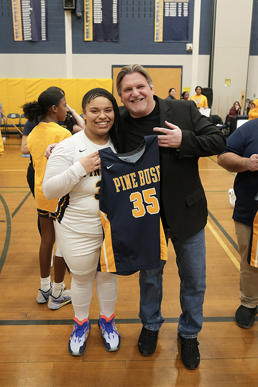 A man stands on the right smiling and a high school girl in a white basketball uniform. they are both holding a side of a blue and gold basketball jersey that says Pine Bush 35