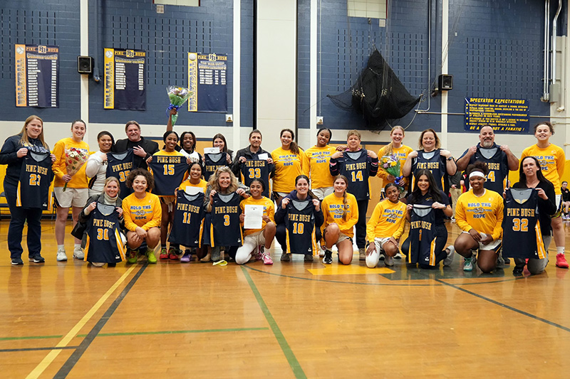 A group of 14 girls high school basketball players with an adult next to each. half are kneeling on the floor and the rest standing behind them. All are wearing gold or blue jerseys.