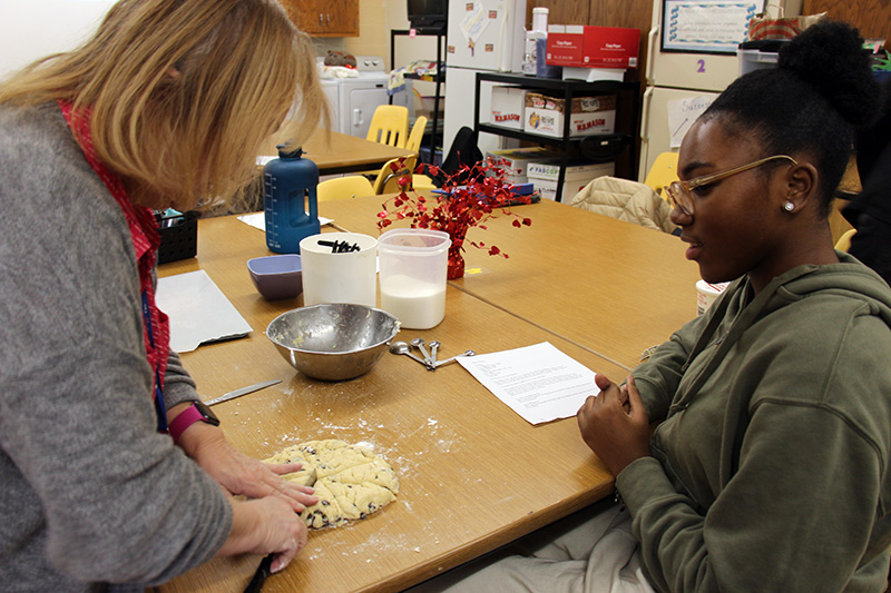 A woman on th eleft uses a knife to cut dough sitting on a table. A young woman on the right watches.