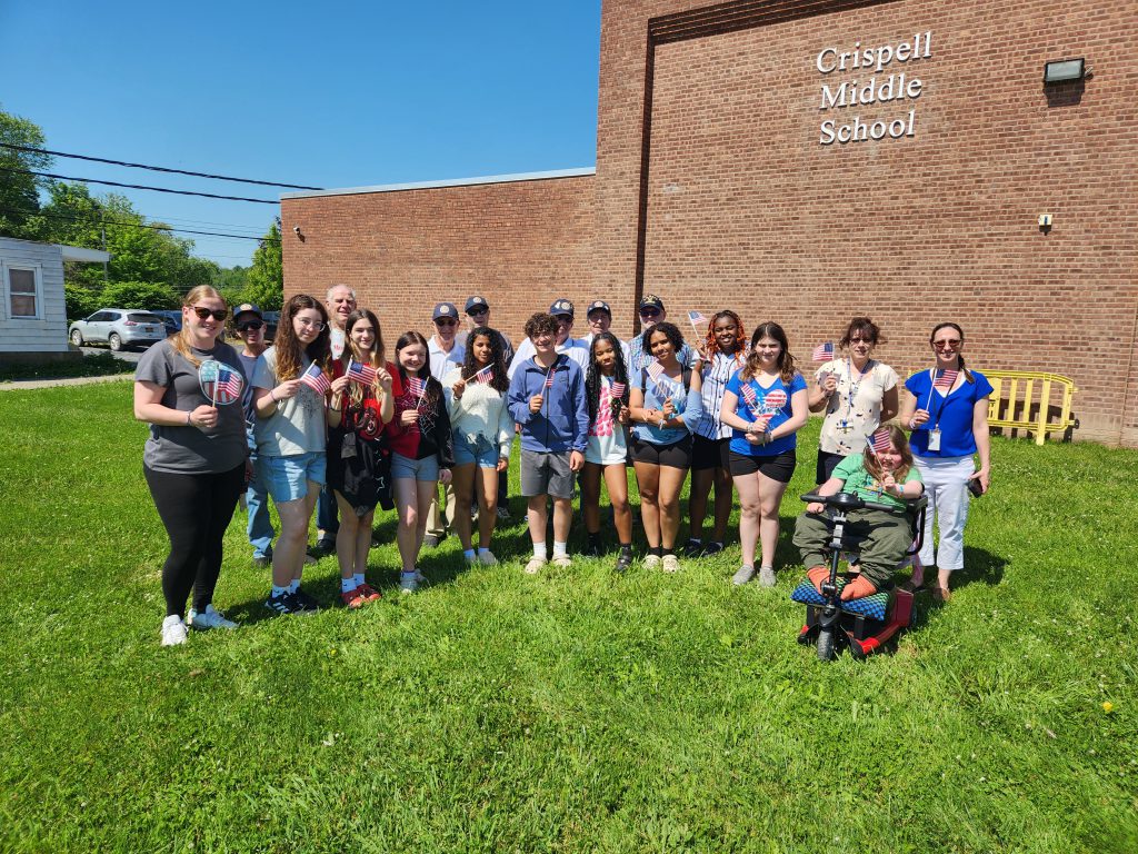 A group of about 10 middle school students with 10 adults. All of the students and some adults ar eholding small America flags. There is a brick building in the background that says Crispell Middle School. 