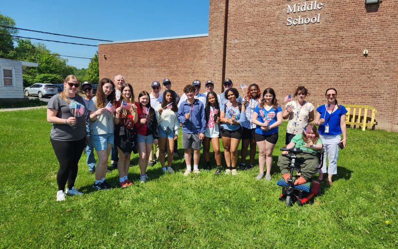A group of about 10 middle school students with 10 adults. All of the students and some adults ar eholding small America flags. There is a brick building in the background that says Crispell Middle School.