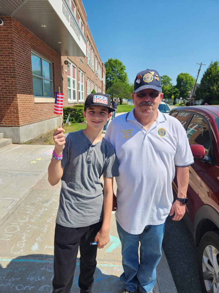 A man stands on the right wearing a blue hat with a veterans insignia on it. A middle school boy stands on the left holding up a flag.