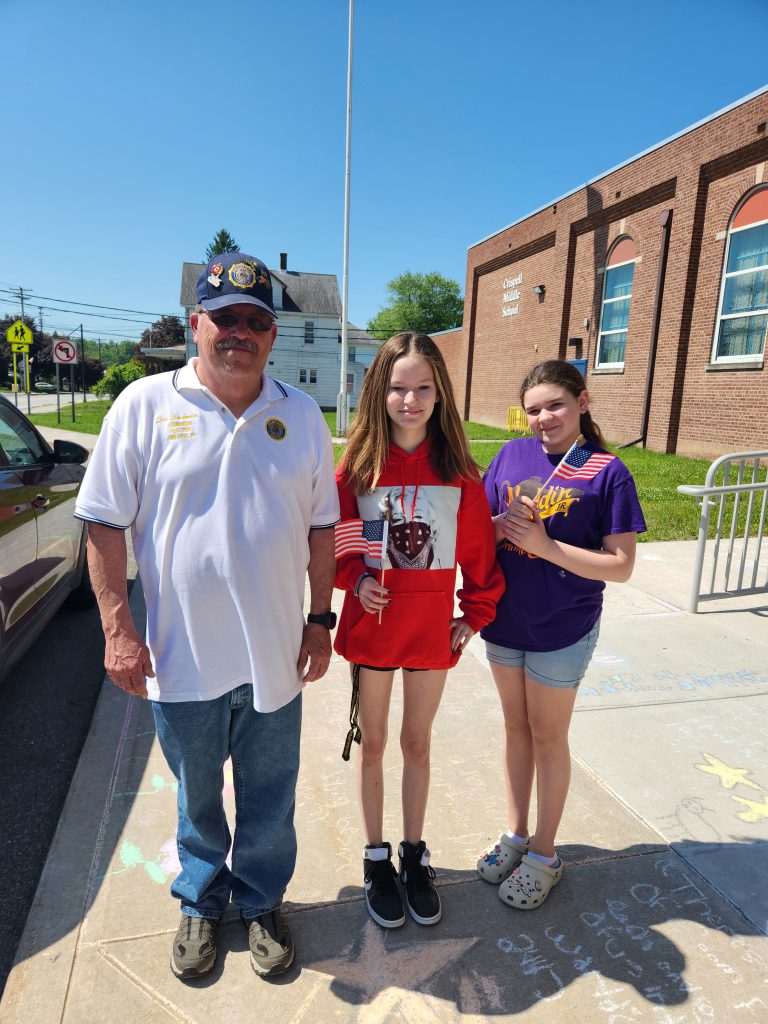 Two middle school girls stand with a man who is wearing a veterans cap. The girls are holding flags.