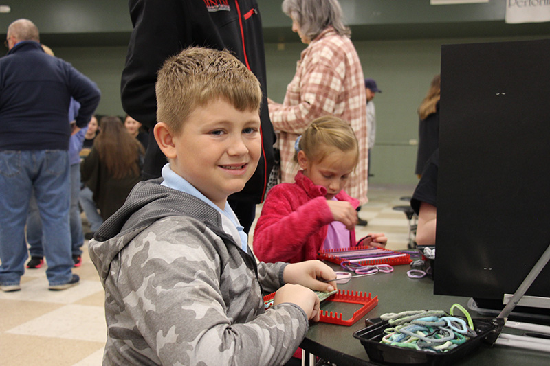 A boy with short blonde hair sits at a table weaving a pot holder.