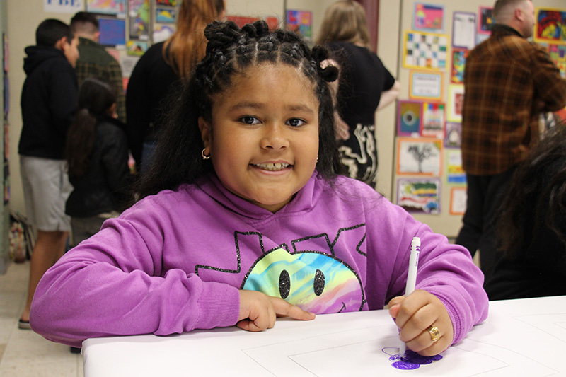 A girl wearing a purple shirt with a big smiley face on it. She is smiling and has the front of her hair in braids.