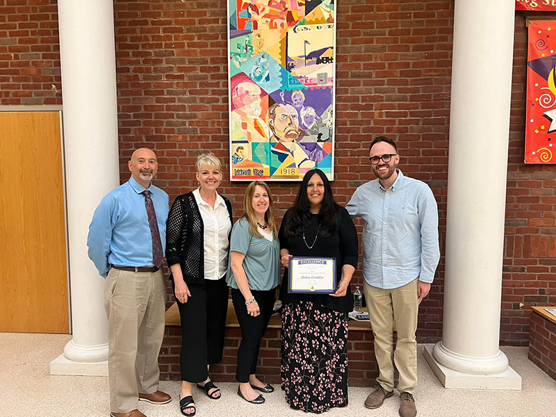 Five adults stand together with a large piece of artwork on the wall in the back. The woman second from right is holding a certificate.