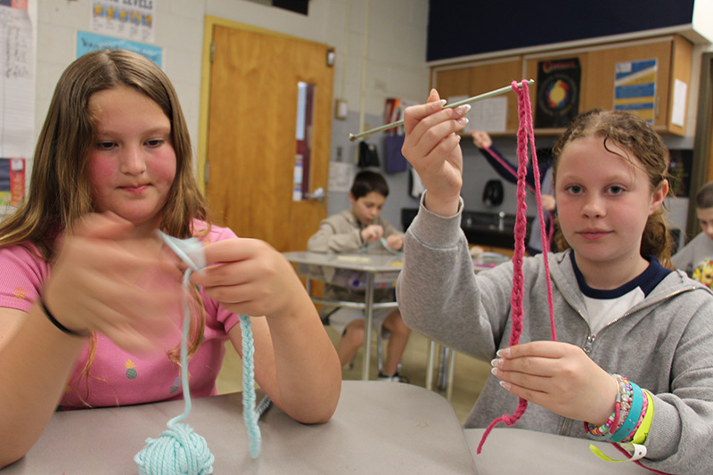 Two fifth-grade boys work on making yarn dolls. A woman with long dark hair and glasses sits between them helping.