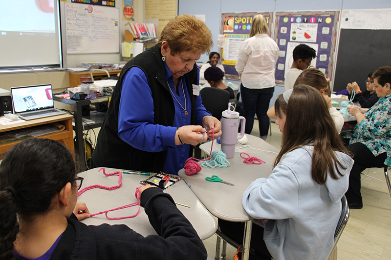 A woman wearing a blue shirt and black vest helps a fifth-grade student with her crochet.