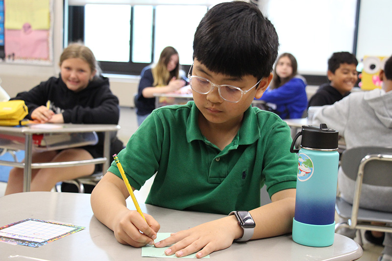 A fifth-grade boy with short dark hair, wearing a green shirt and glasses draws on a piece of paper while sitting at his desk.