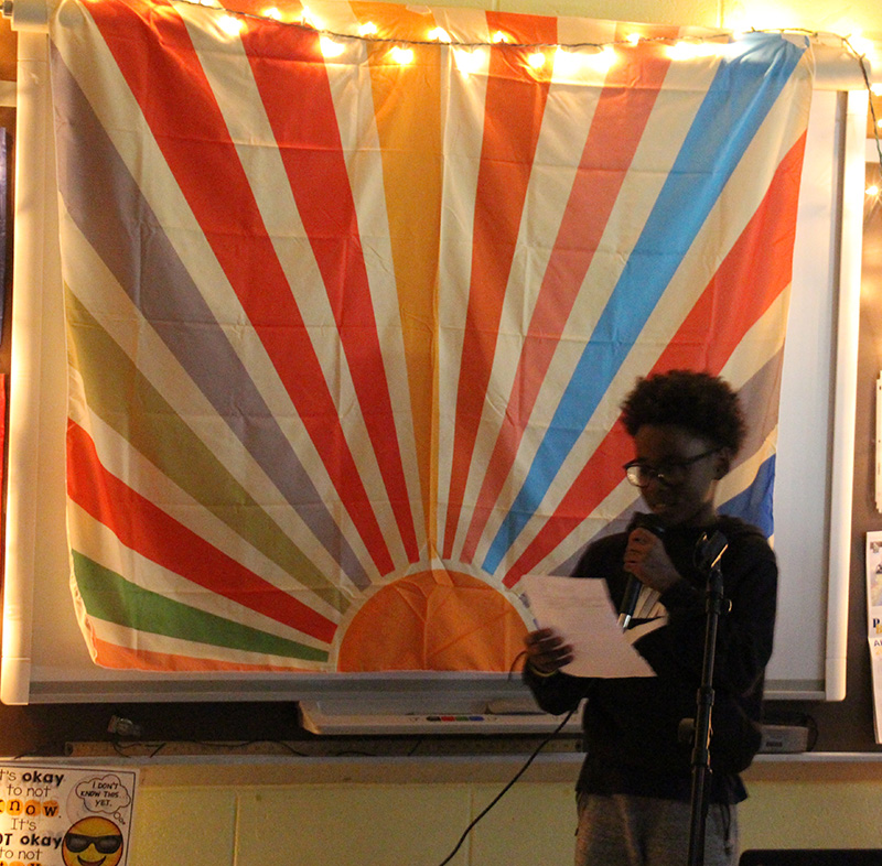 A backdrop of a sun with multi color rays. There is a fourth-grade boy standing in front reading a poem.