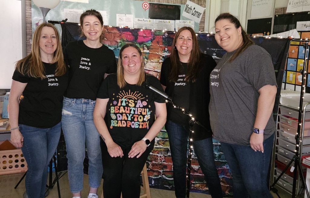 Five adults stand together smiling. They all have shirts on that talk about poetry.