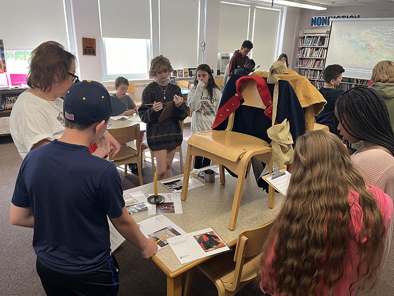 Several middle school students stand around a table looking at Revolutionary War artifacts.