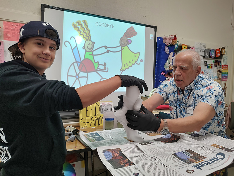 An older man on the right, wearing a blue printed short-sleeve shirt holds a cast while a middle school student holds the top of it. They are smiling.