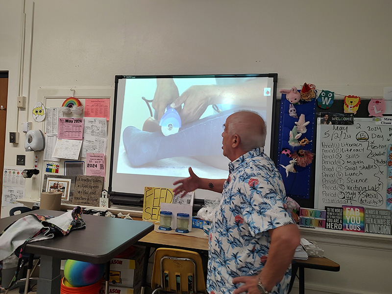 An older man in a blue print shirt gives a presentation to a class. He is gesturing to a screen with an informational slide on it.