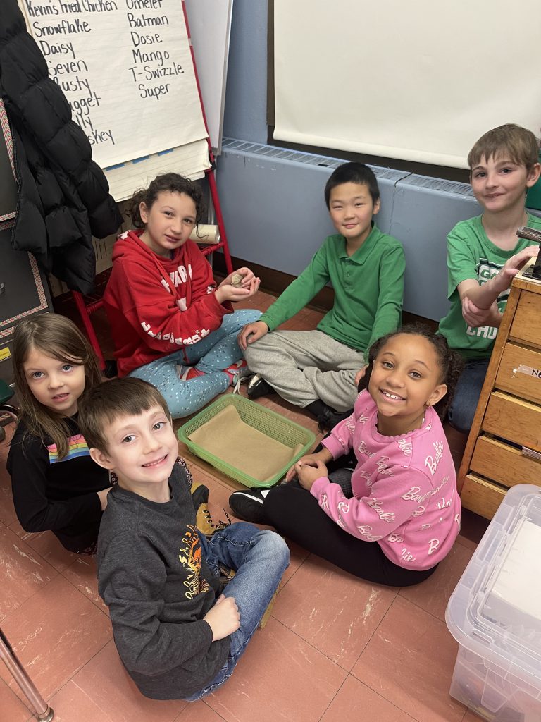 Six fourth-grade students sit on a floor with baby chicks. The kids are all looking at the camera smiling.