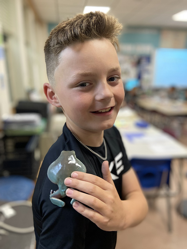 A boy smiles. He is holding a shiny frog on his shoulder.