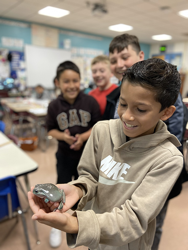 a boy with dark hair wearing a light tan hoodie smiles as he holds a shiny frog in his hands. Several kids behind him are looking and smiling.