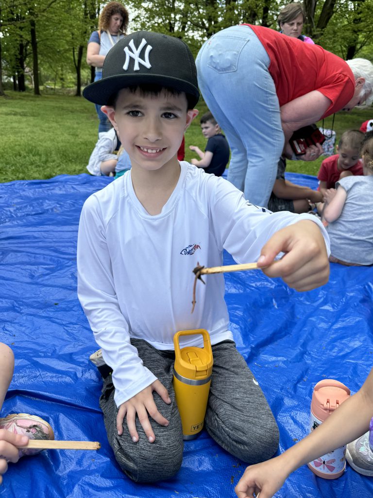 A kindergarten kid wearing a NY Yankees ballcap and white shirt smiles as he holds a worm on a stick.