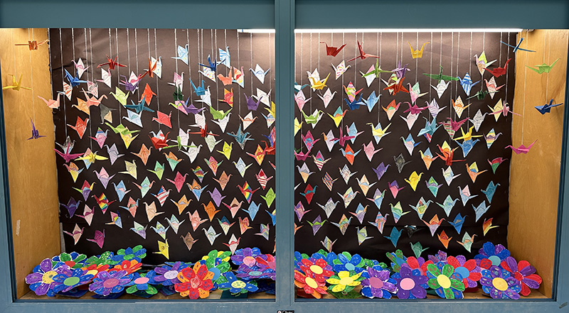 A glass display case with lots of colorful tiny paper cranes hanging in it.