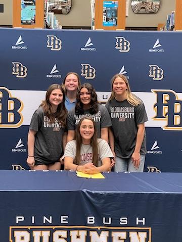 A high school senior sits at a table with four high school students standing behind her. They are all smiling.