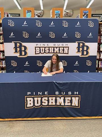 A high school senior sits at a table with a tablecloth that says Pine Bush Bushmen. She is signing a piece of paper.