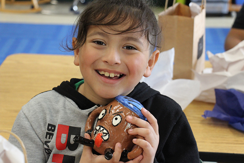 An elementary age student smiles broadly holding up a clay figure.