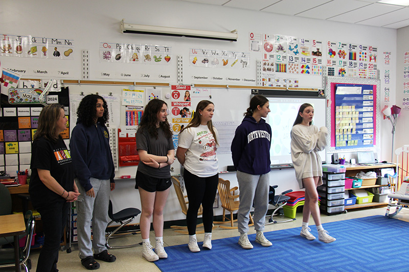 Six young women stand at the front of a classroom. One is speaking to the class.