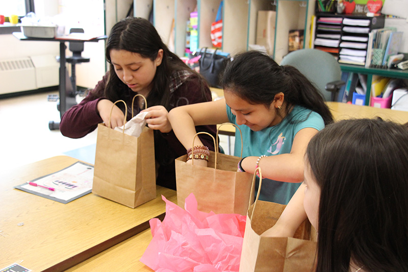 Two elementary girls sit at a table and look eagerly into brown gift bags.