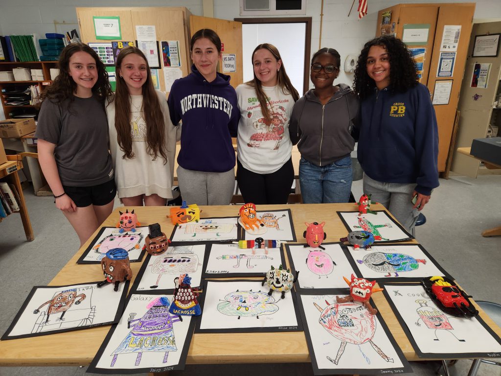 Six high school girls stand in a row with a table filled with artwork in front of them.