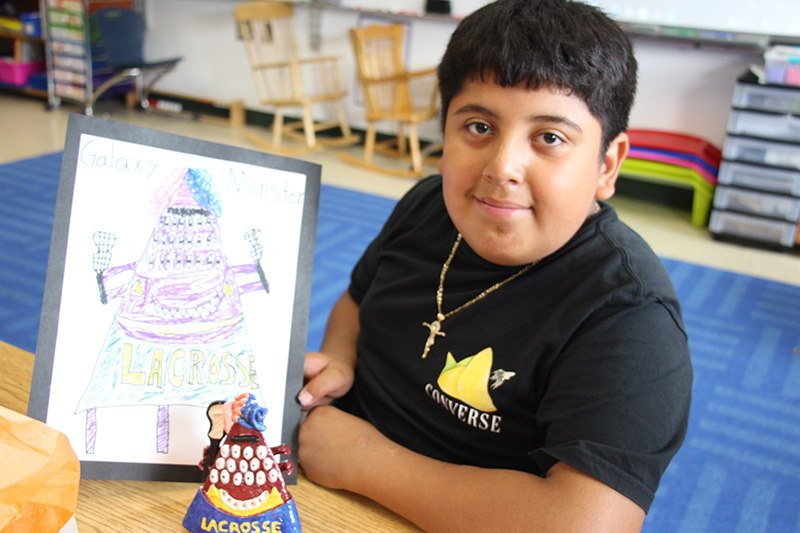 An elementary age boy with short dark hair holds up a drawing he made. In front of it is a clay figure of that drawing.