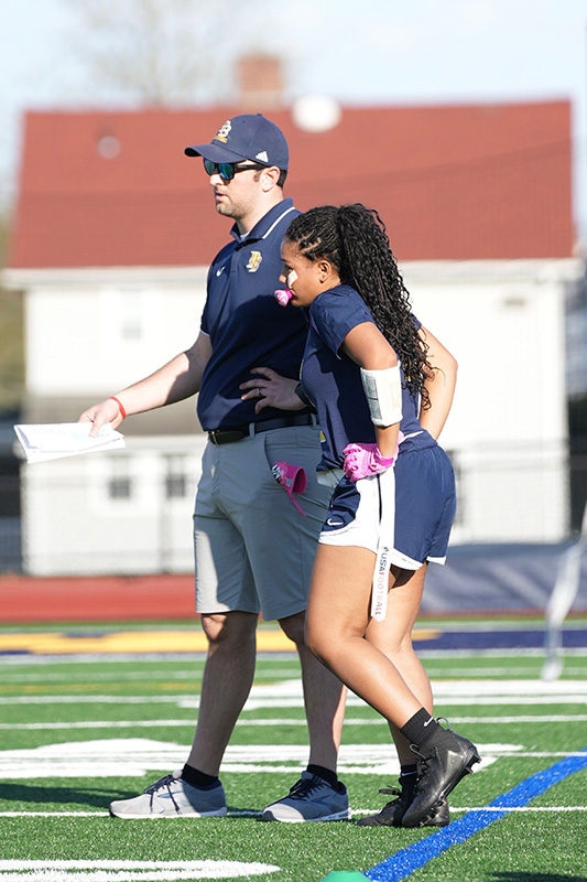 A tall man wearing sunglasses, a blue baseball type cap and blue shirt stands on the sidelines with a high school girl in a navy blue flag football uniform. She has long dark hair.