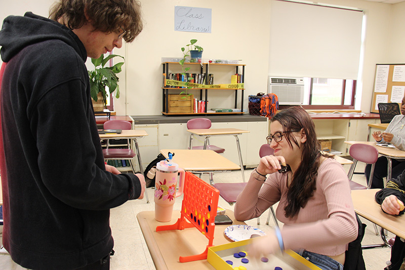 A high school girl sits at a desk playing connect four with a woman standing nearby.