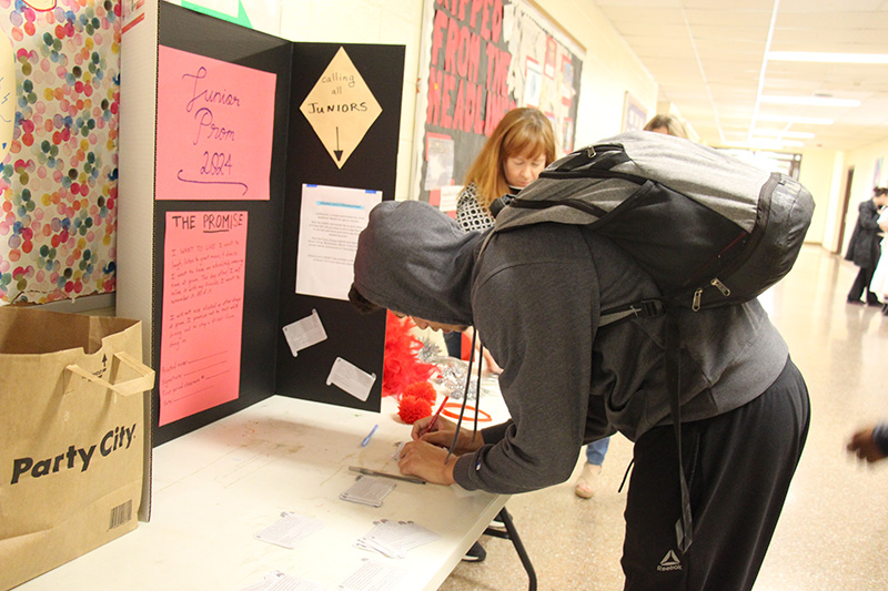 A high school student in a gray hoodie leans on a table and signs a piece of paper.