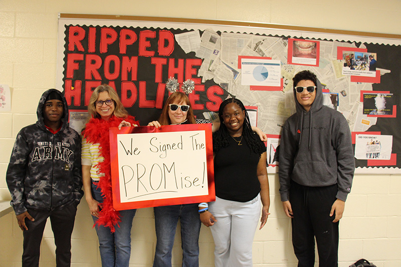 Two adults and three high school students stand together and hold a sign that says We signed the PROMise! They are smiling.