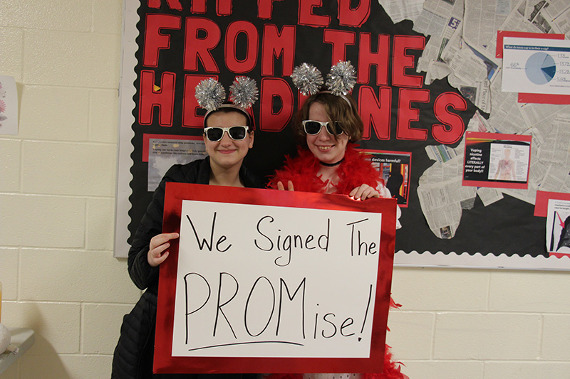 Two students with glittery headbands and red boas, and sunglasses stand smiling holding a sign that says We signed the PROMise!