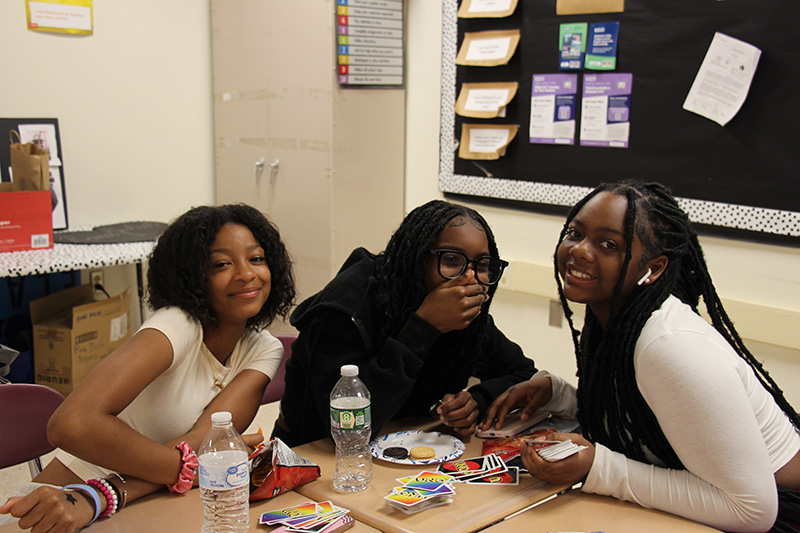 Three high school girls sit at a table smiling and eating snacks.