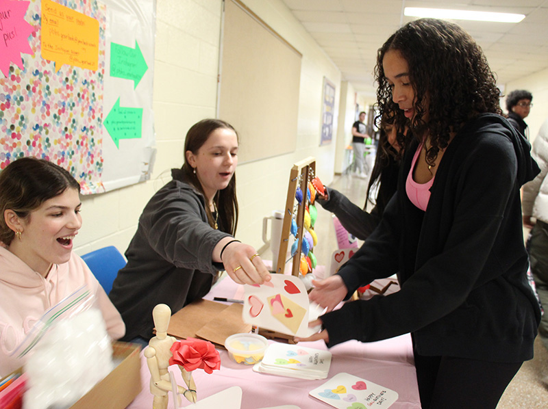 Two high school girls sit at a table with small pieces of artwork they are selling. One girl is reading a handmade card with hearts on it.