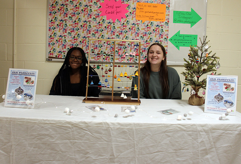 Two high school girls are sitting at a table with handmade ornaments. They are smiling.