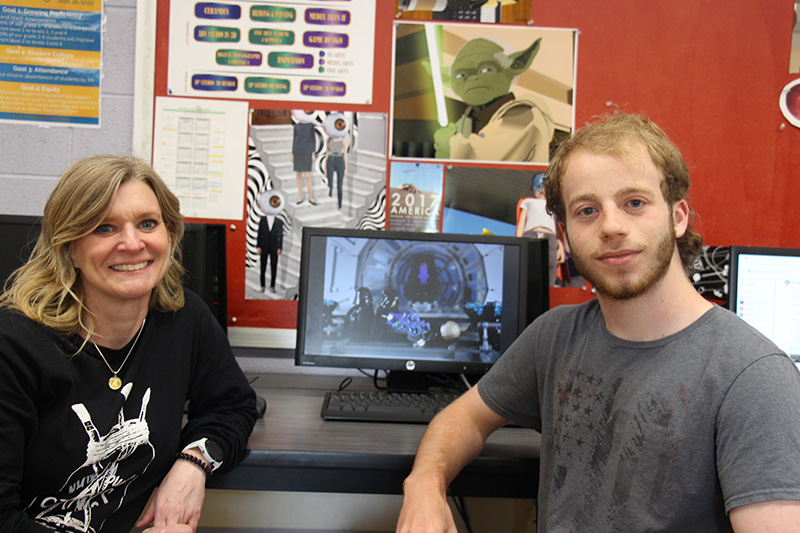 A woman with shoulder length blond hair sits on the left and a high school students sits on the right. There is a laptop in the center with artwork on the screen.