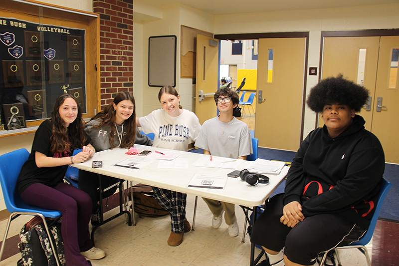 A group of five high school students smile as they sit around a table that has papers on it.